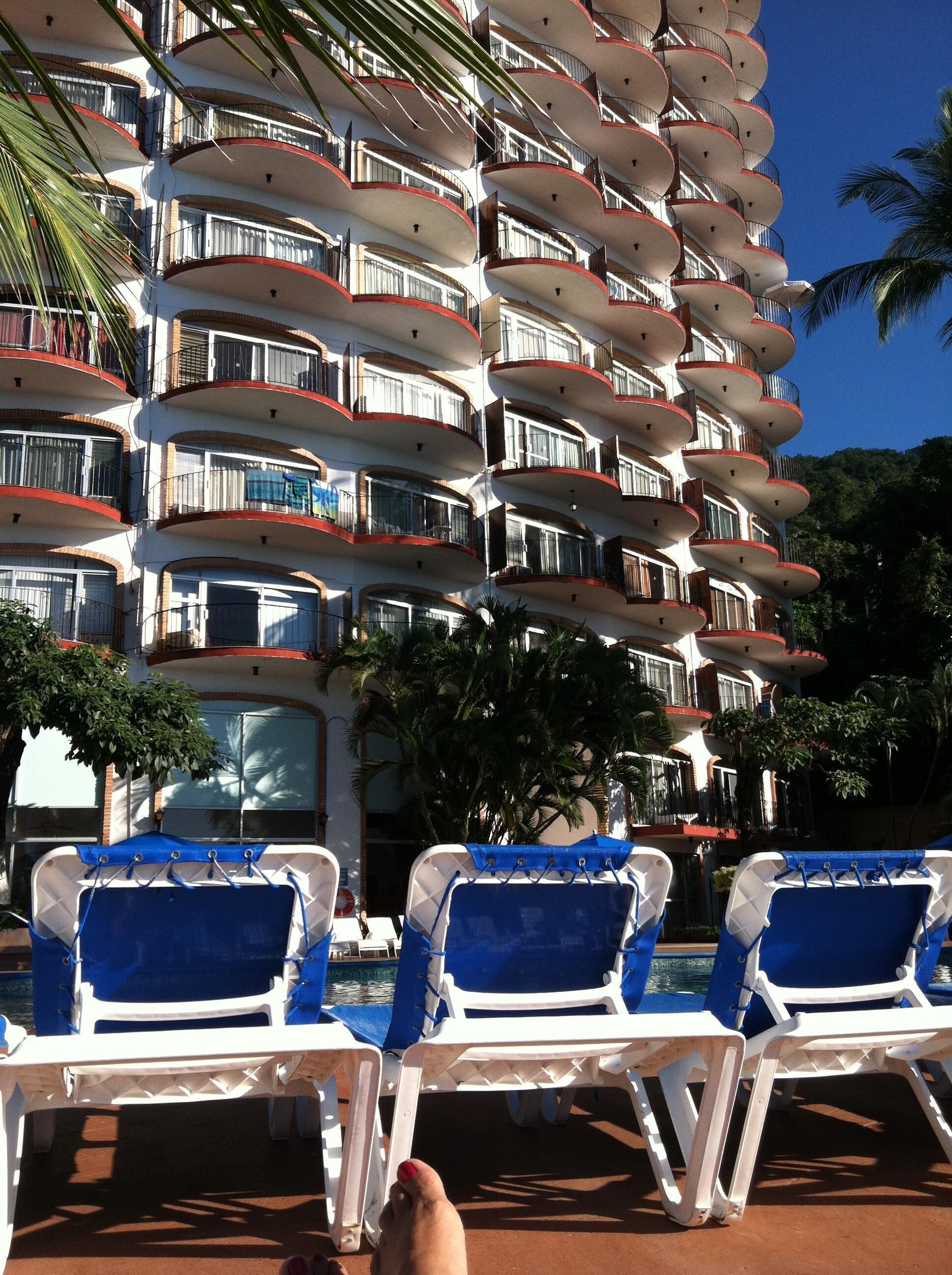 Un hôtel à plusieurs étages doté de balcons, d’un espace piscine avec des chaises longues et de palmiers.