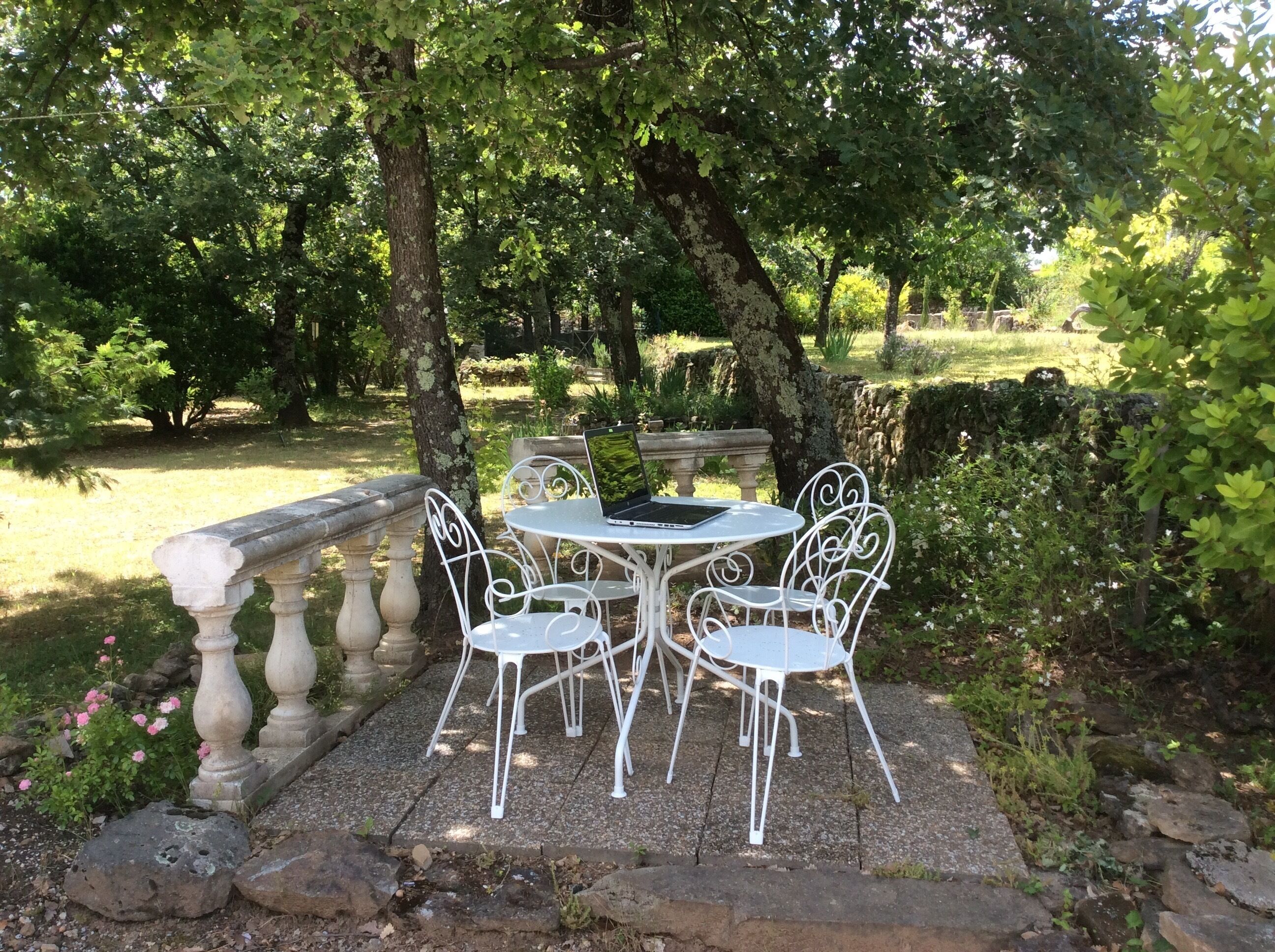 Une terrasse de jardin avec une table ronde et quatre chaises blanches, entourée de balustrades en pierre et d’une végétation luxuriante.