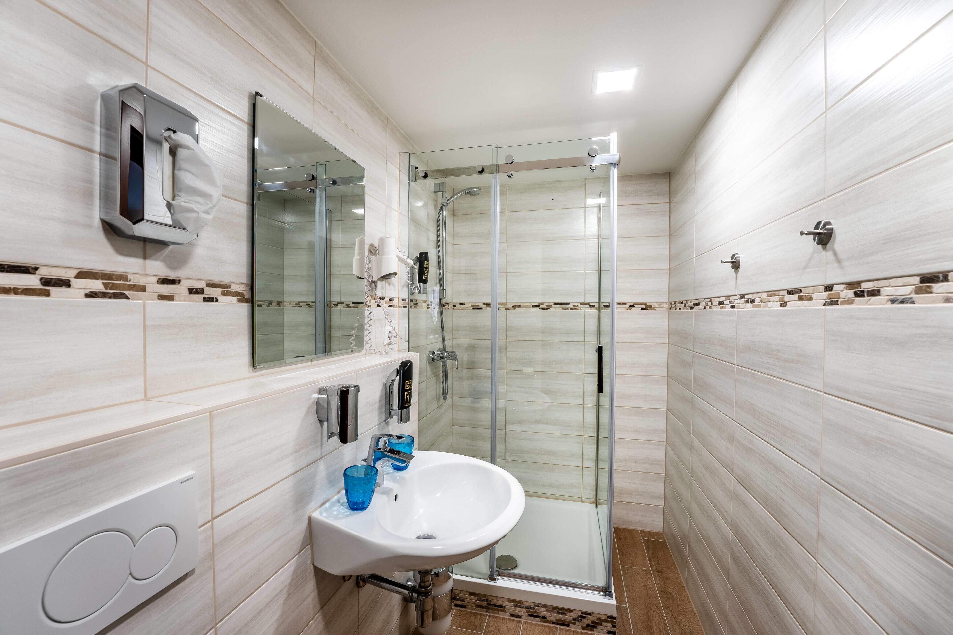 A modern bathroom with a glass shower enclosure, a white sink with blue glasses, and a towel dispenser.