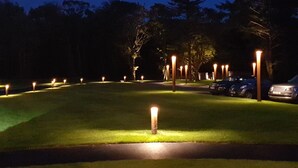 Een buitengebied 's nachts, met gelijkmatig verdeelde lichtjes langs een gebogen pad, omgeven door bomen en geparkeerde auto's.