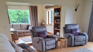 Een moderne woonkamer met een grijze bank, twee relaxfauteuils, een houten salontafel en een boekenkast.