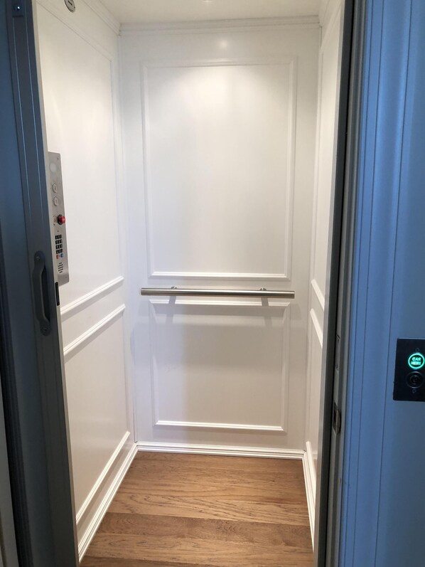 A narrow, enclosed elevator with a wooden floor, a metal handrail, and a control panel with a red button.