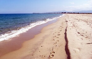 En sandstrand med fotspår som leder ner mot vattnet, en klar himmel och avlägsna berg.