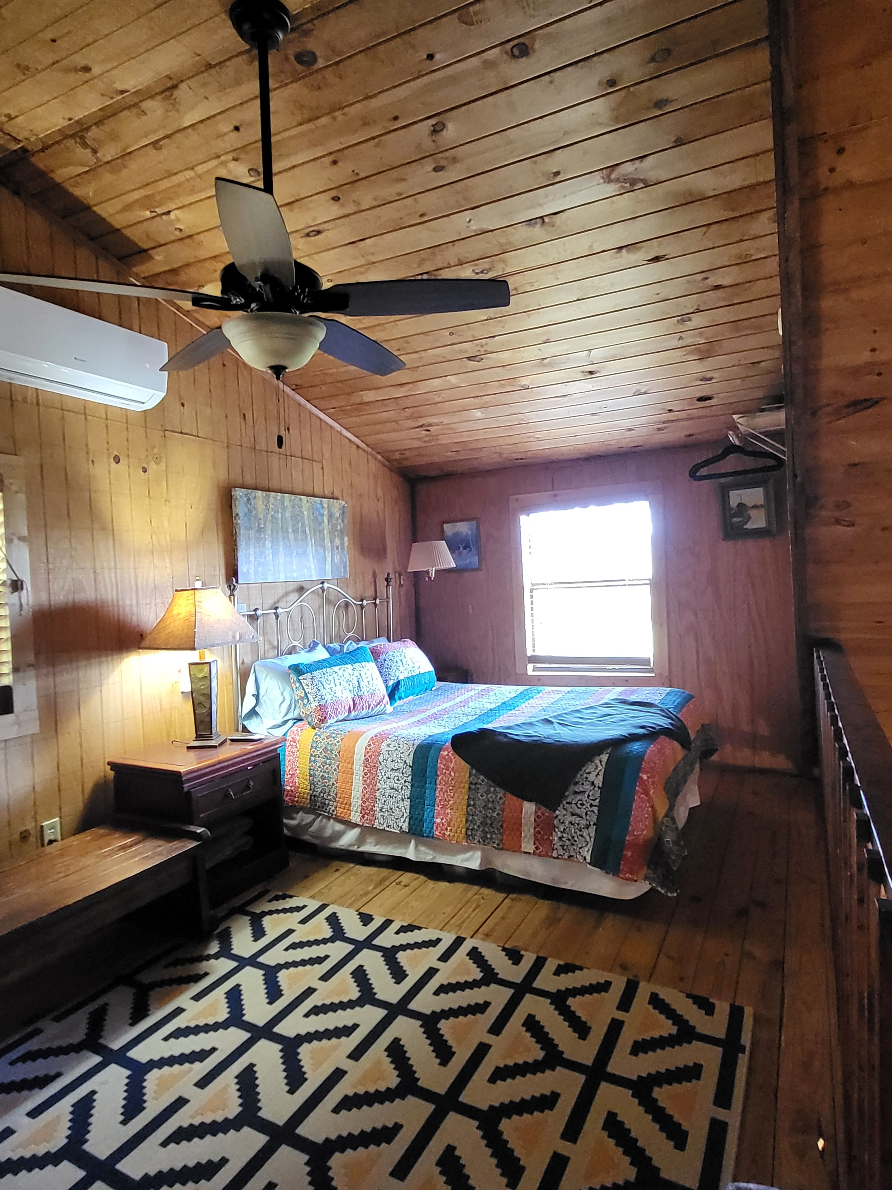 Une pièce avec un plafond en bois, un lit recouvert d’une couverture aux couleurs vives, un bureau avec une lampe et un tapis à motifs.