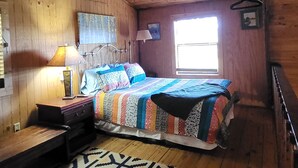 Una habitación con techo de madera, una cama con colcha colorida, un escritorio con lámpara y una alfombra estampada.