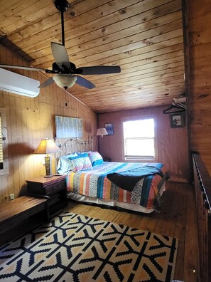 Une pièce avec un plafond en bois, un lit recouvert d’une couverture aux couleurs vives, un bureau avec une lampe et un tapis à motifs.