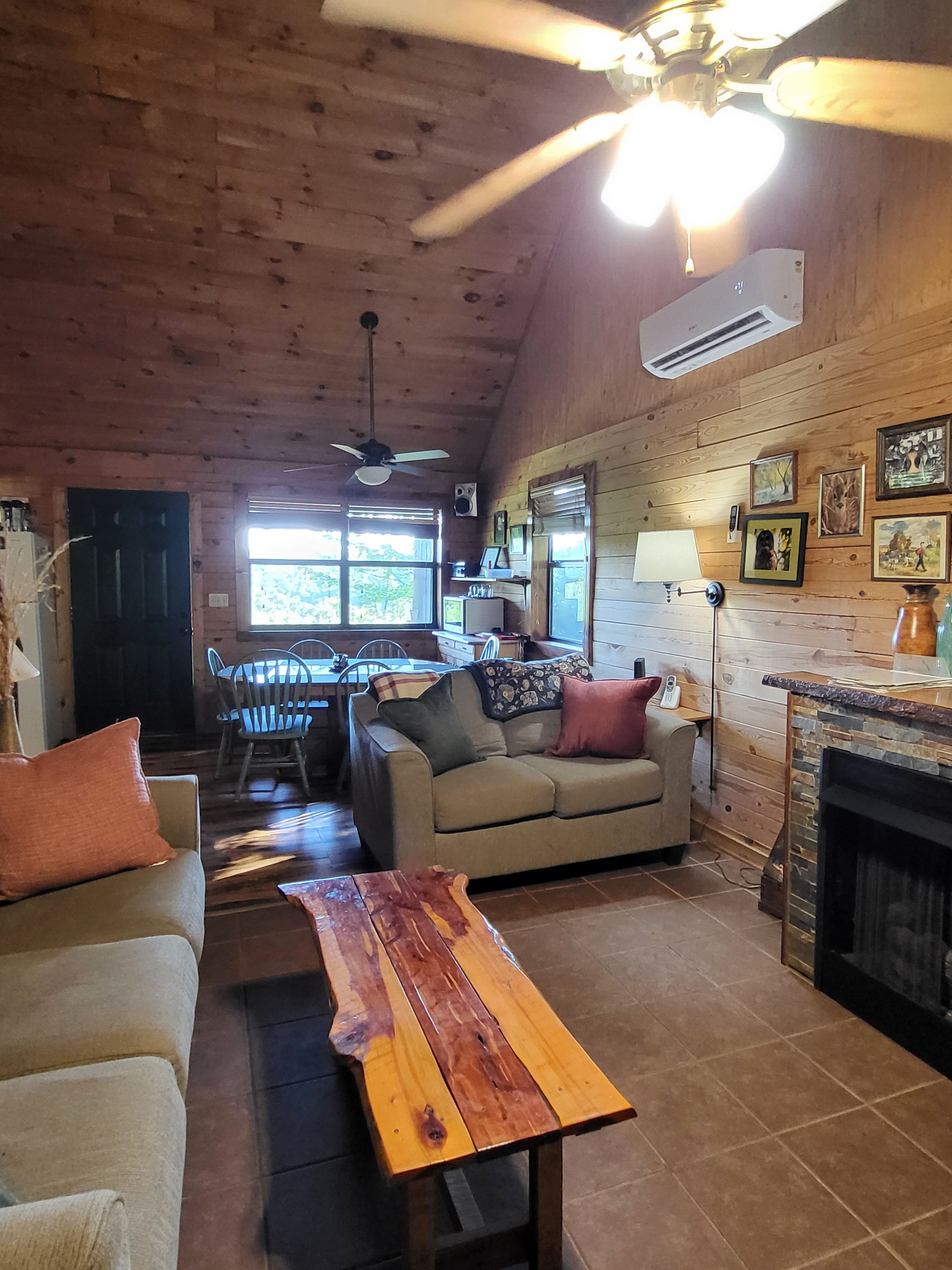 A cozy cabin interior with a wooden ceiling, a dining area, a sofa, a fireplace, and a ceiling fan.