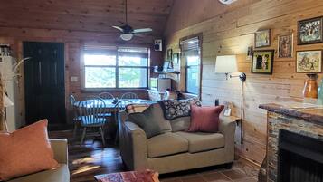 A cozy cabin interior with a wooden ceiling, a dining area, a sofa, a fireplace, and a ceiling fan.