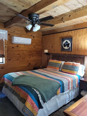 A wooden-paneled room with a bed, a ceiling fan, an air conditioning unit, a framed picture, and a bedside lamp.