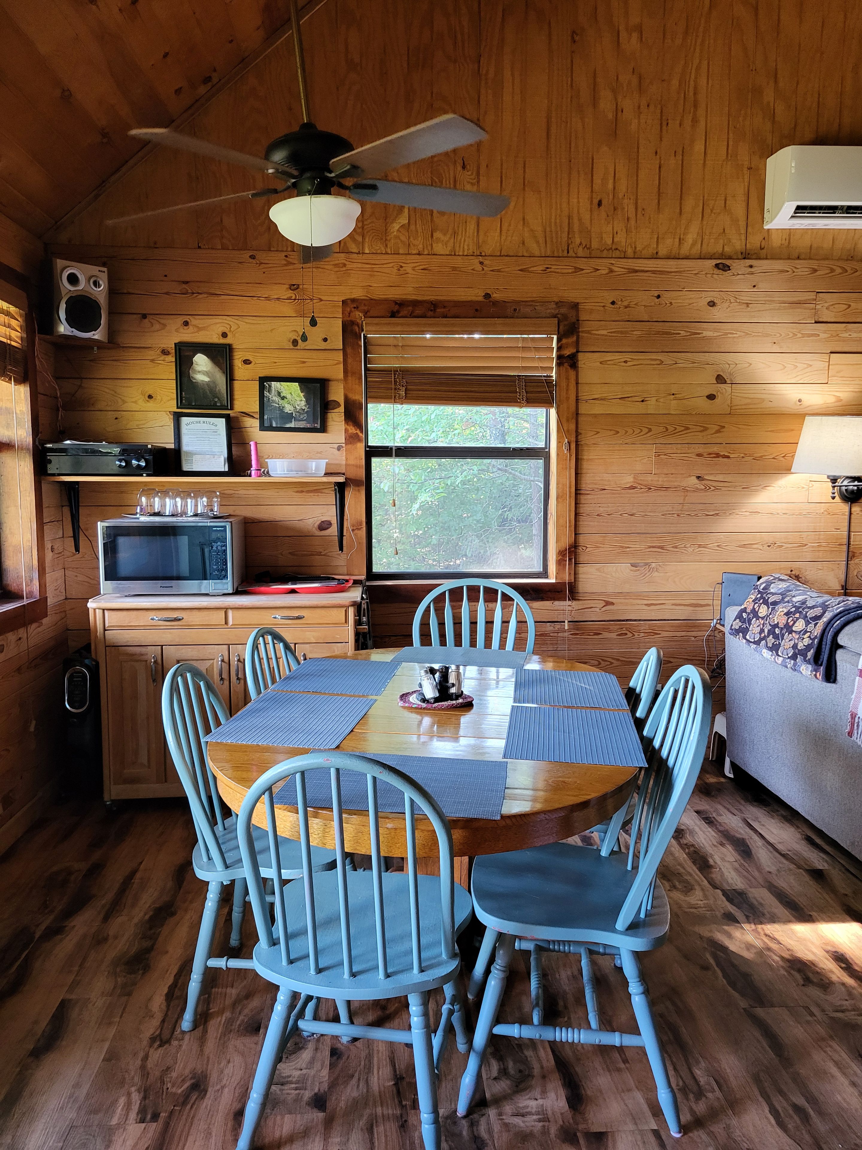 A wooden cabin interior with a dining table, chairs, a ceiling fan, a microwave, and a window with blinds.