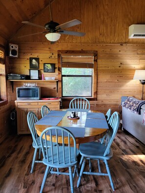 A wooden cabin interior with a dining table, chairs, a ceiling fan, a microwave, and a window with blinds.