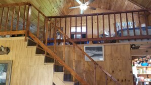 Interior de madera con una escalera que sube a una zona superior, un ventilador de techo y un cuadro en la pared.