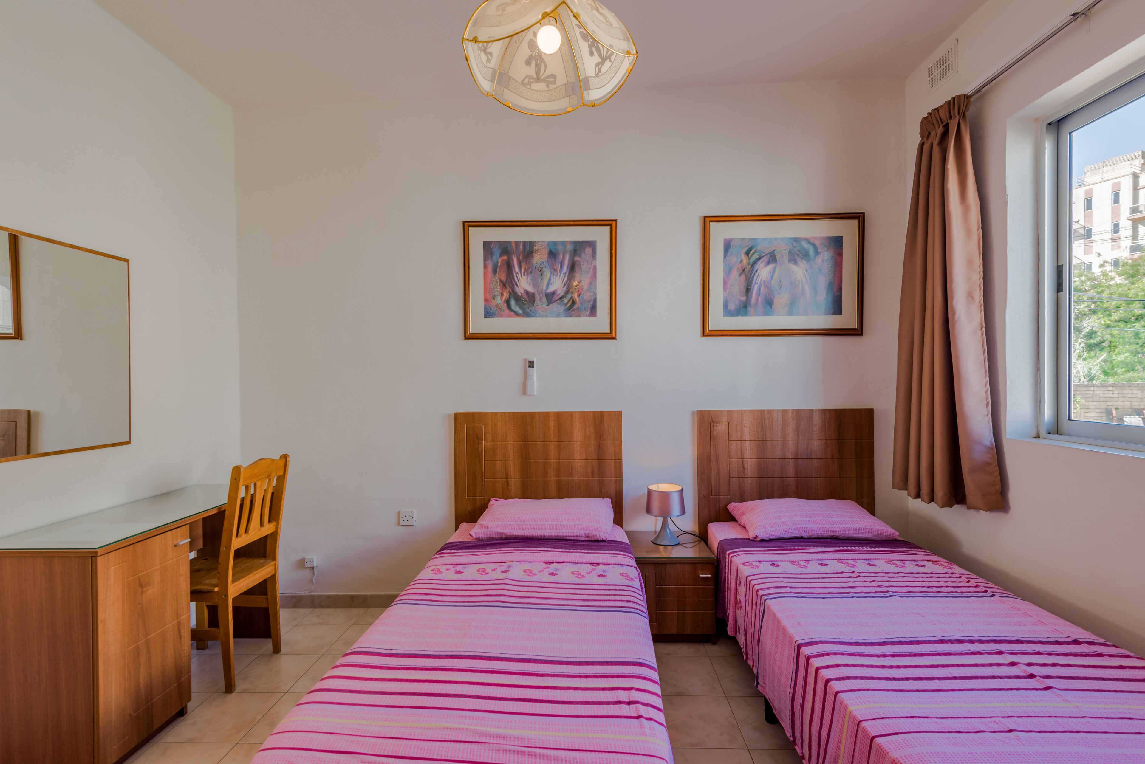 A hotel room with two beds, a desk, and a chair. There are two framed artworks on the wall, a window with curtains, and a ceiling light fixture.