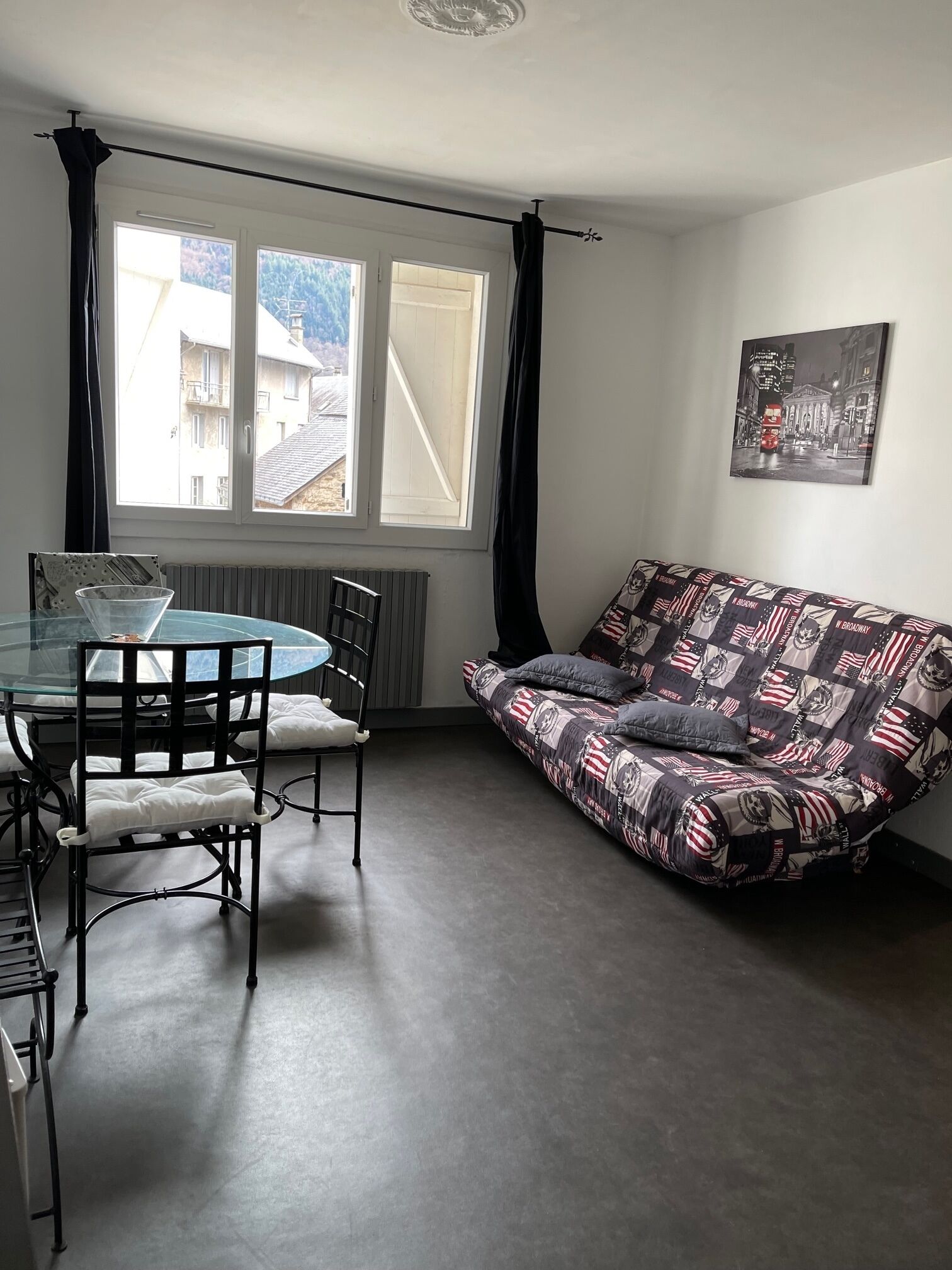 Un salon avec une table à manger à plateau de verre, deux chaises, un canapé à motifs et une vue sur les immeubles par la fenêtre.