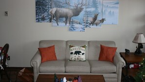 A living room with a grey sofa, two orange pillows, a 'Welcome to the Cabin' pillow, and a coffee table with figurines.