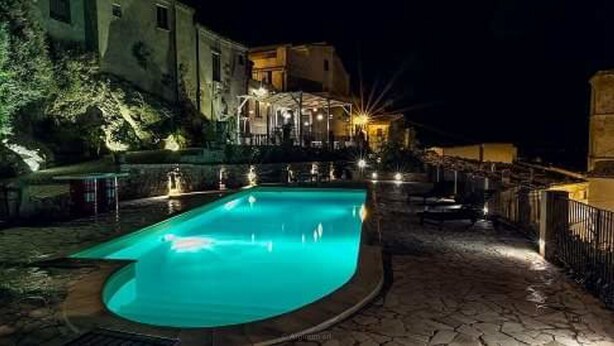 A nighttime swimming pool with a stone deck, surrounded by buildings and outdoor lighting. - Case al Borgo - Albergo diffuso (Agira)