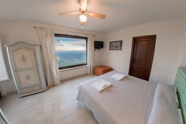 A bedroom with a bed, a window with a view, a ceiling fan, a wardrobe, and a door. - Case al Borgo - Albergo diffuso (Agira)