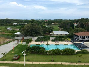 A large outdoor swimming pool with a surrounding grassy area and a few palm trees. There are buildings in the background and a clear sky. - Refreshing and relaxing beach apartment (San José)