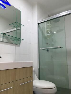 A modern bathroom with a glass shower enclosure, a white toilet, and a wooden vanity with a mirror and glass shelves. - Refreshing and relaxing beach apartment (San José)