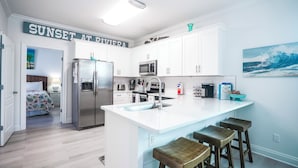 Una cocina moderna con armarios blancos, un refrigerador de acero inoxidable, un microondas y un lavamanos. Hay un cartel sobre el lavamanos que dice "SUNSET AT RIVIERA".