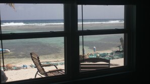 Vista del océano a través de una ventana, con una silla y una estructura pequeña visibles en la playa.
