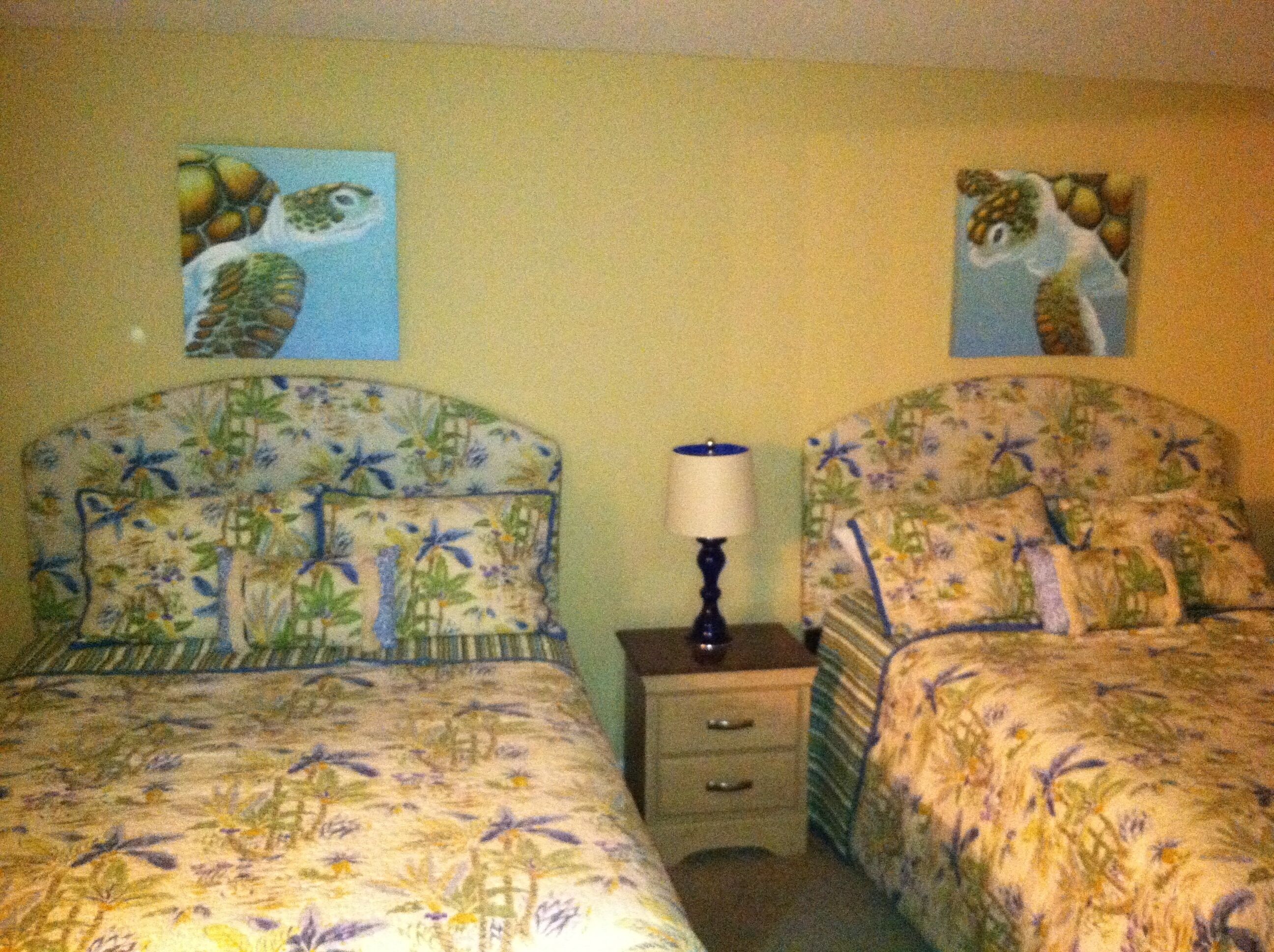 Un dormitorio con dos camas, cada una con ropa de cama floral, una mesita de noche con lámpara, y dos cuadros decorativos en la pared.