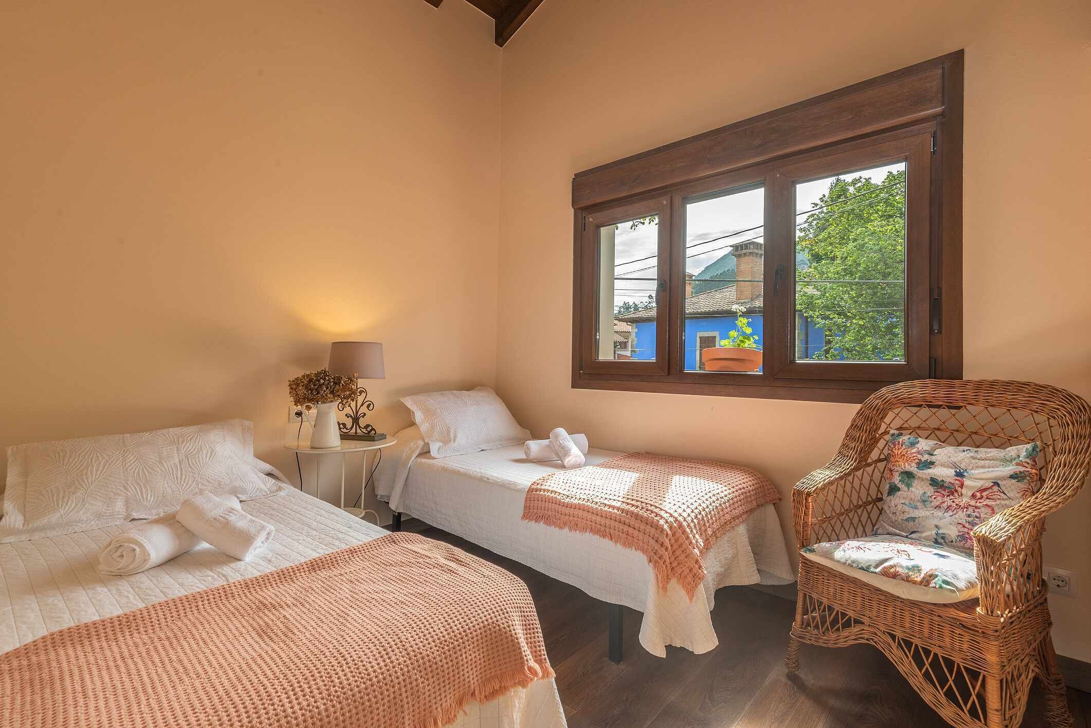 A bedroom with two beds, a wicker chair, and a window with a view of the outside.