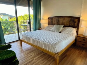 A bedroom with a wooden bed, two bedside tables with lamps, a balcony with chairs, and a view of greenery. - OCEAN VIEW, WALK TO BEACH, SLEEPS 8, POOL & HOT TUB, 3BDR + PRIVATE MASTER SUITE (Kihei)