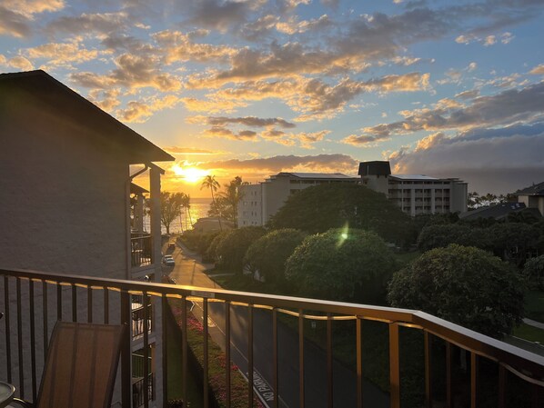 A balcony with a view of a sunset over a cityscape, including buildings and palm trees. - OCEAN VIEW, WALK TO BEACH, SLEEPS 8, POOL & HOT TUB, 3BDR + PRIVATE MASTER SUITE (Kihei)
