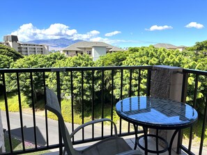 A balcony with a glass table and chairs, overlooking a lush garden and a road. - OCEAN VIEW, WALK TO BEACH, SLEEPS 8, POOL & HOT TUB, 3BDR + PRIVATE MASTER SUITE (Kihei)