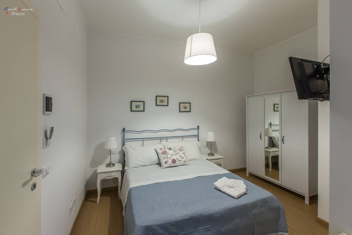 Un dormitorio con cama, mesitas de noche, un espejo y un televisor montado en la pared.