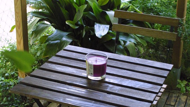 Une table en bois sur laquelle est posée une bougie dans un récipient en verre, le tout entouré d’une végétation luxuriante.