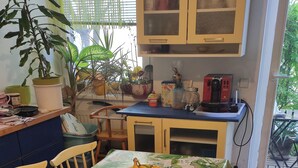 Une cuisine avec une table à manger recouverte d’une nappe à motifs floraux, des chaises, un saladier rempli de fruits et divers appareils de cuisine.