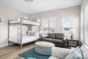 A modern bedroom with a bunk bed, a sofa, a round ottoman, and a view of the outdoors through the windows.