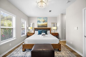 A bedroom with a large bed, two bedside tables with lamps, a bench, a painting on the wall, and a window with blinds.
