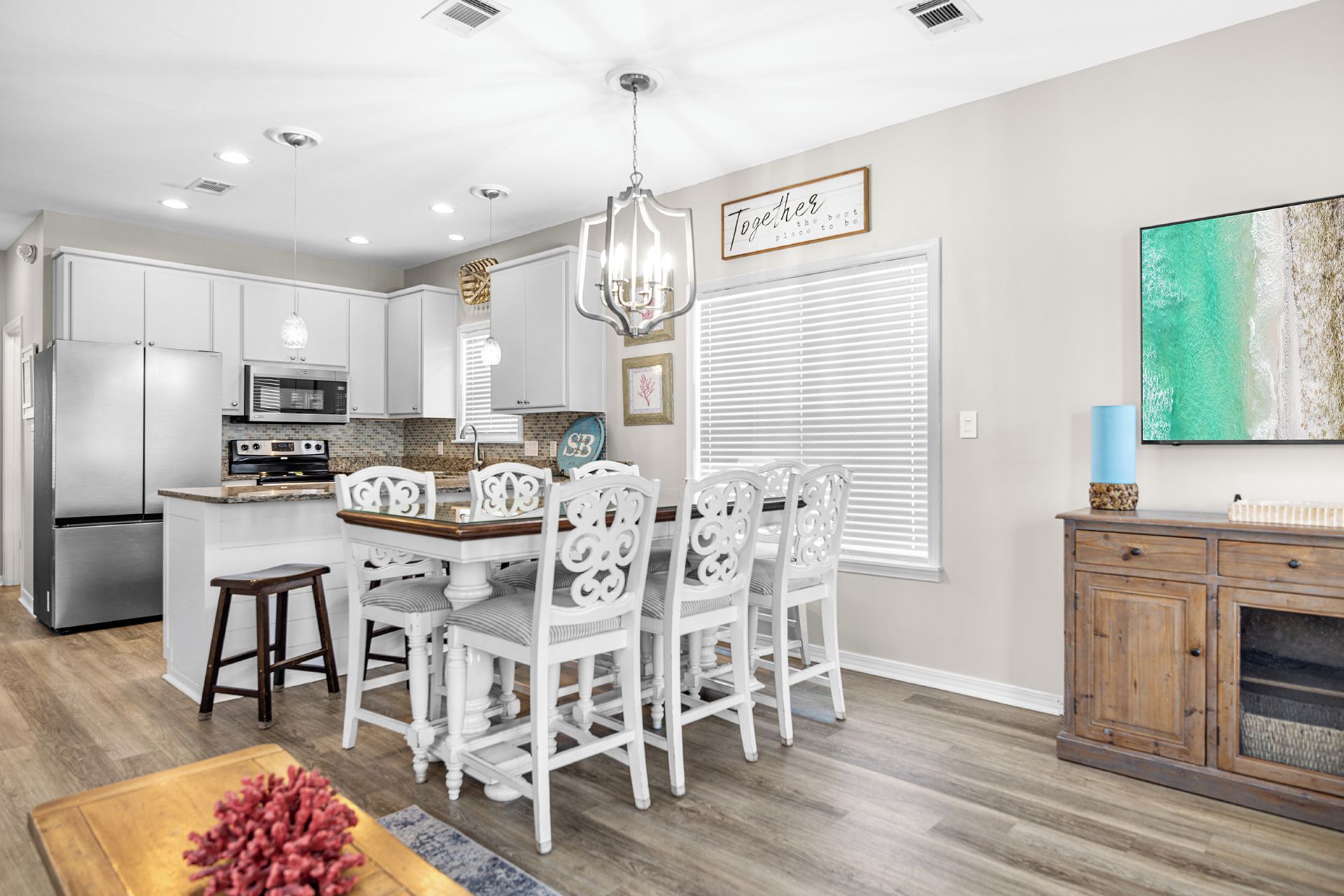 A modern kitchen with white cabinetry, a dining area with white chairs, and a large window with blinds.