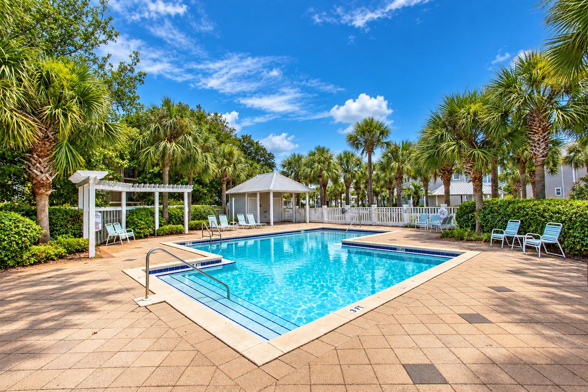 A swimming pool with lounge chairs and a covered seating area, surrounded by palm trees and a clear sky.
