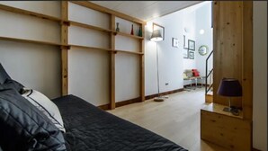 Quarto com cama, estantes de madeira, abajur e uma mesinha.