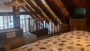 Ein Zimmer mit schrägem Dach, Holzbalken, einem Bett mit gemustertem Überwurf, einem Fernseher auf einer Holzkommode und einem dekorativen Totempfahl.