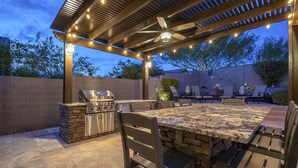 Un patio al aire libre con una estación de barbacoa de piedra, una mesa con sillas y luces colgantes.