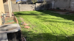 Un patio trasero con césped cuidado, un sendero de ladrillos y una unidad de aire acondicionado grande.