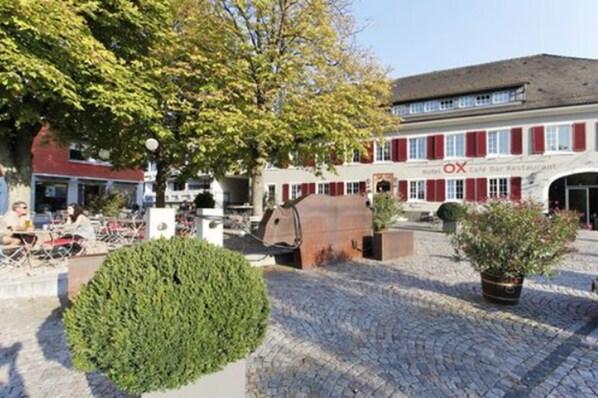 Un hotel con terraza exterior, un árbol grande y un edificio con contraventanas rojas.