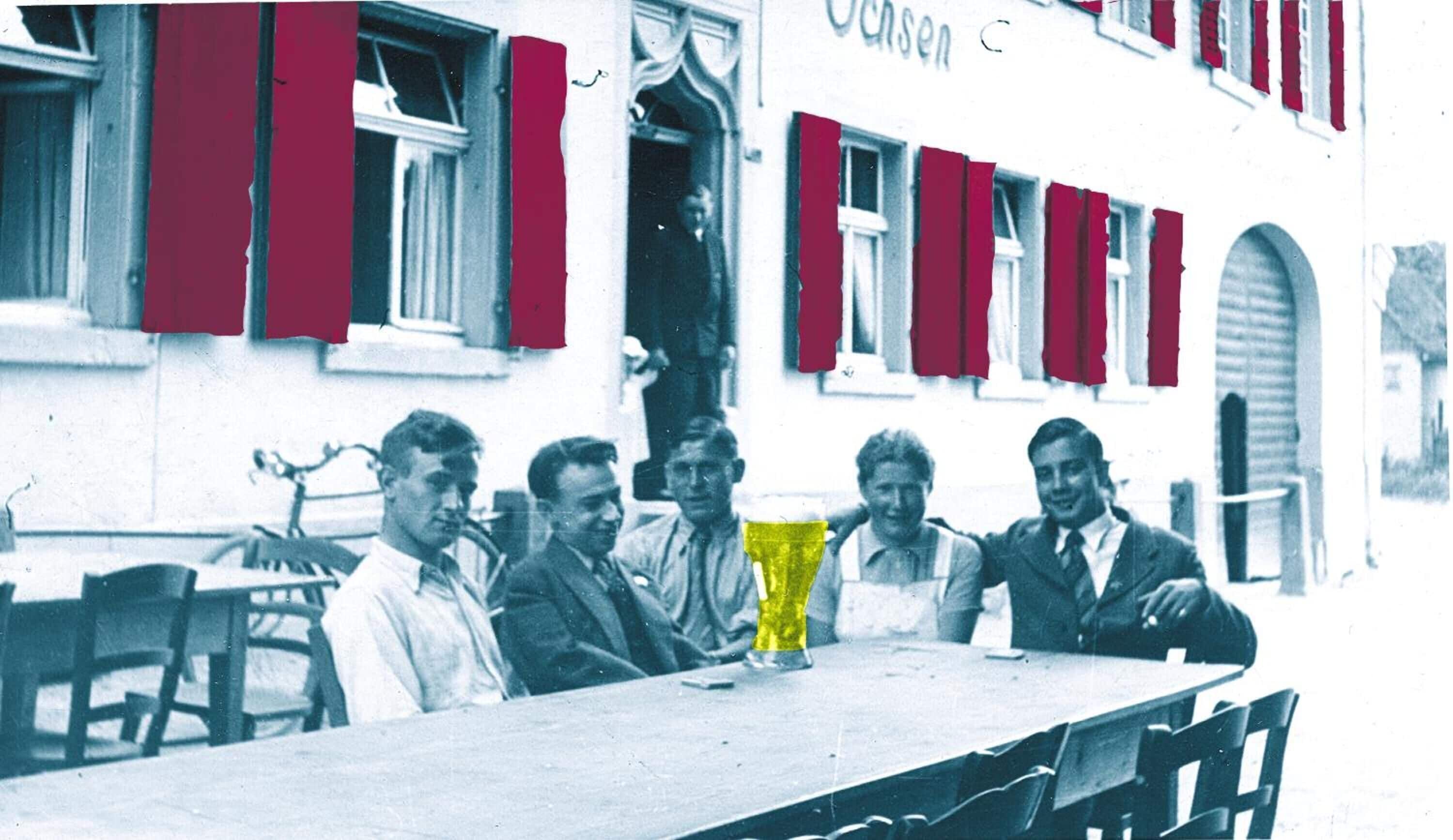 Un groupe d’hommes est assis à une table autour d’un objet jaune placé au centre, devant un bâtiment aux volets rouges sur lequel on peut lire l’inscription « Casein ».