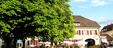 Ein Café im Freien mit Tischen und Stühlen, umgeben von Grünflächen und Gebäuden.