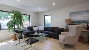 Una sala de estar moderna con un sofá de cuero, una mesa de centro de vidrio, un sillón blanco y un ventanal con vistas a árboles.
