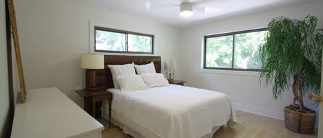 Un dormitorio con una cama, mesitas de noche, ventilador de techo, dos ventanas y una planta en maceta.