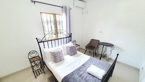 A neatly arranged bedroom with a bed, a chair, a small table, and a window with bars. - Appartement Maképé B.M 1 (Douala)