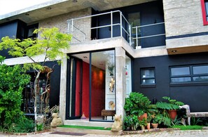 Un edificio moderno con entrada de vidrio, un balcón y diversas plantas en macetas.