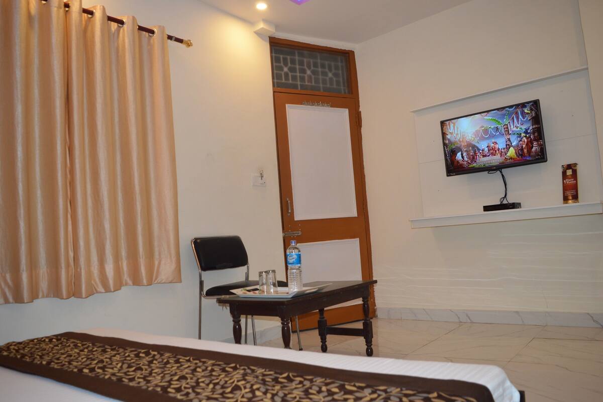 A hotel room with a bed, a small table with a bottle and glasses, a chair, a TV mounted on the wall, and a window with curtains.
