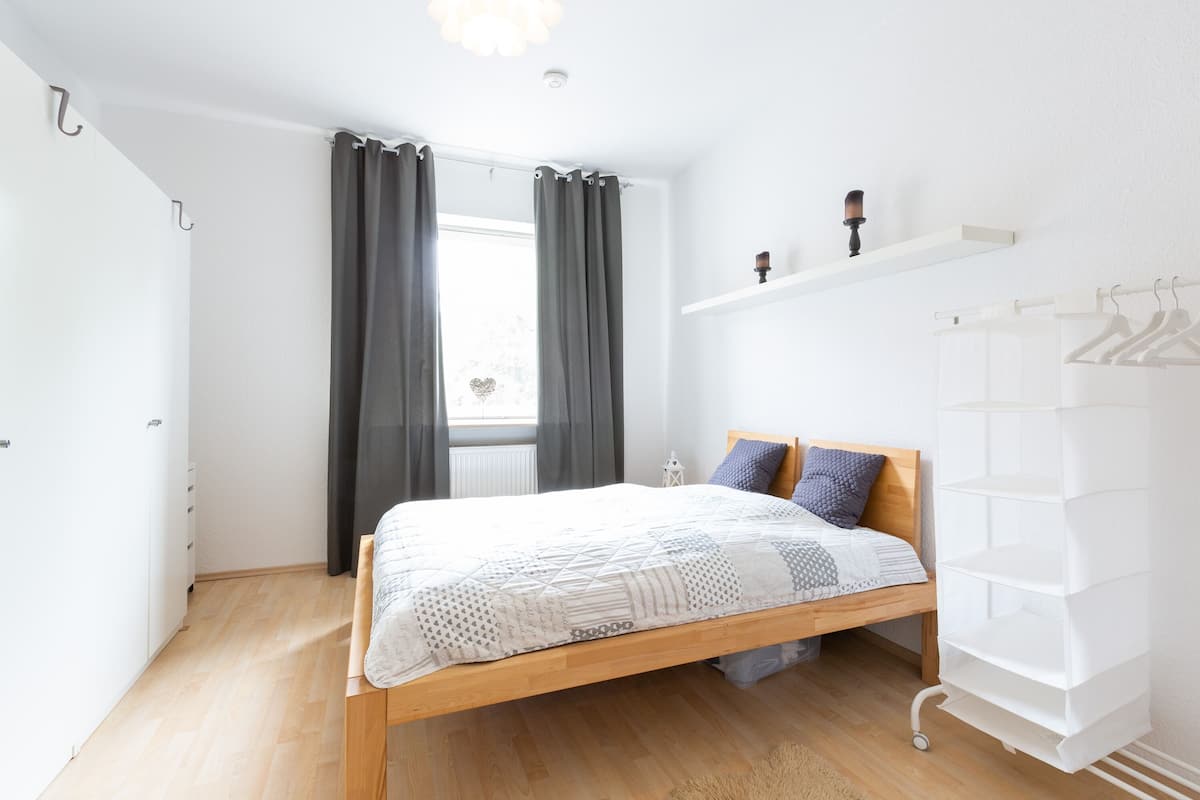 A bedroom with a wooden bed, grey curtains, a white shelf with decorative items, and a white hanging rack.